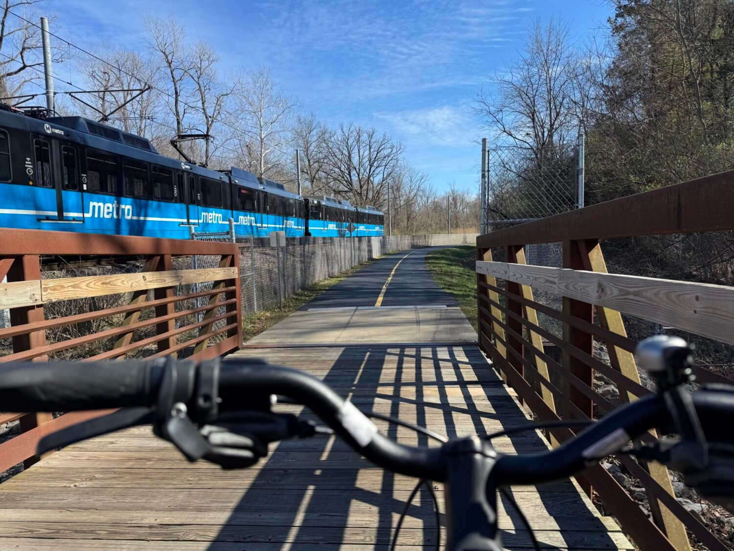 A POV shot of a bicycle riding across a bridge along the MetroBikeLink Trail with the train passing on the left.