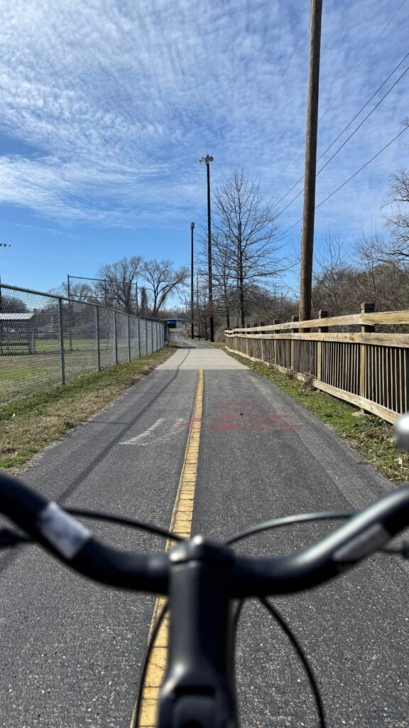 A POV shot of a bicycle riding between a creek bed and baseball field along the Richland Creek Greenway.